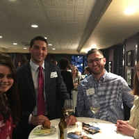 two male alumni and two female alumni gathered at a table with wine and food, smiling for camera